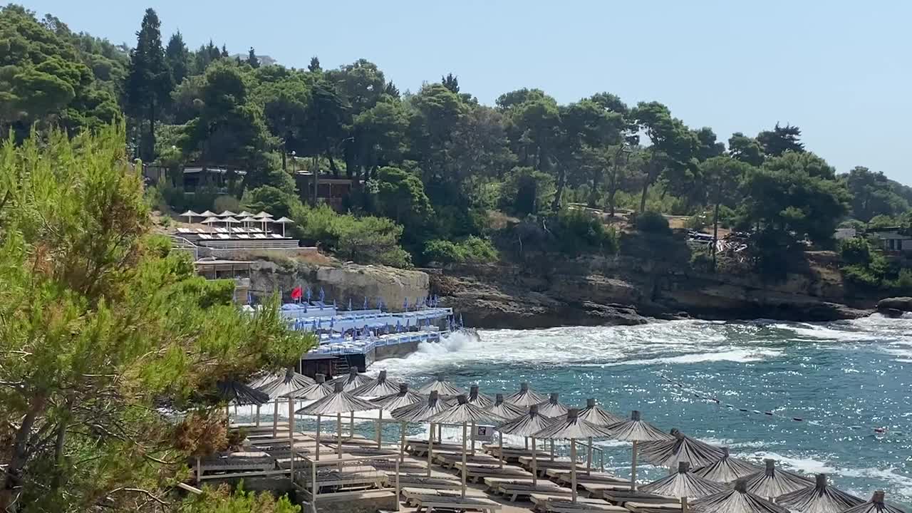 夏天在地中海的天堂海滩度假。阳光明媚的日子里，天堂度假村周围是松林。美丽的蓝色风暴的海浪。休息区的遮阳伞和阳光躺椅，让您放松，放松，放松，乐趣，快乐的假期。视频素材