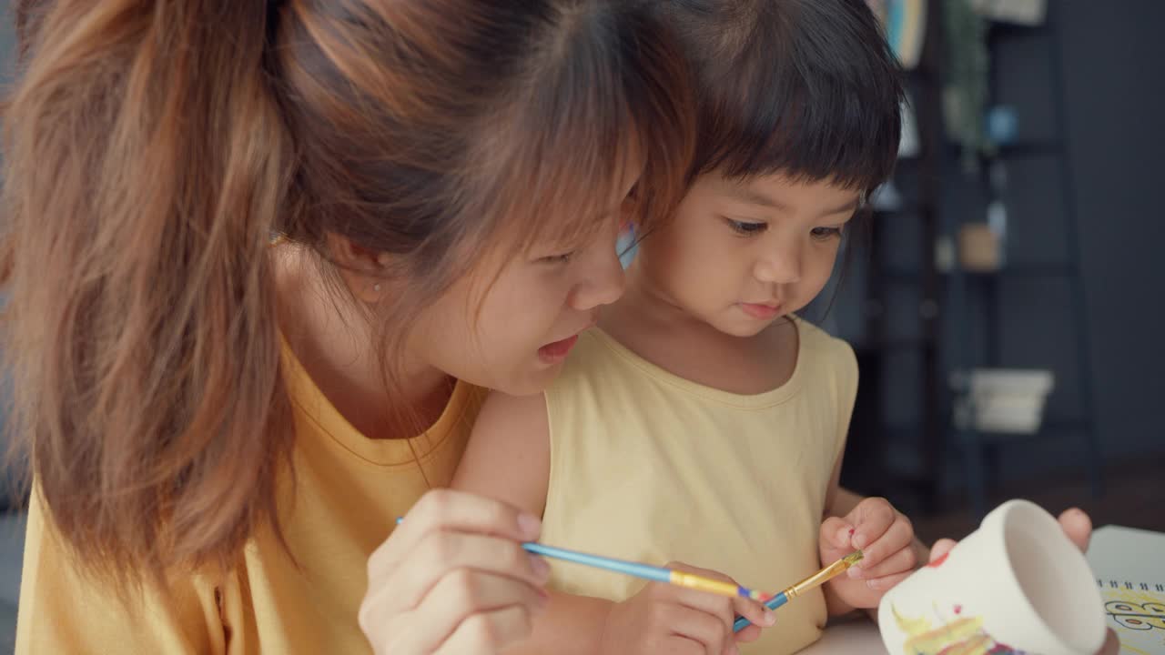 快乐快乐的亚洲家庭妈妈教学步的小女孩在家里客厅的桌子上画画，玩得开心放松。视频素材