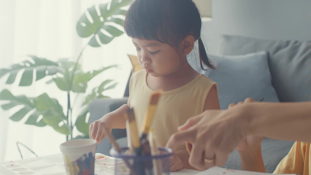 快乐快乐的亚洲家庭妈妈教学步的小女孩在家里客厅的桌子上画画，玩得开心放松。视频素材