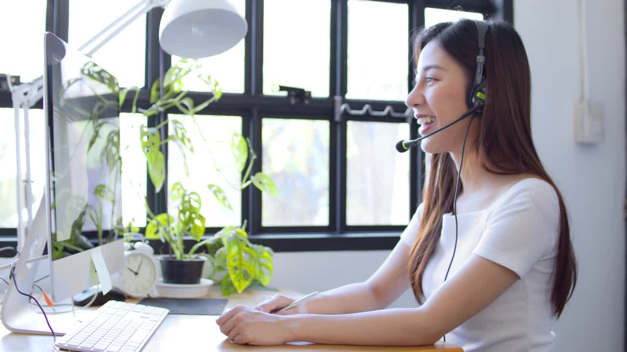 亚洲女孩学生戴着耳机在线学习与skype老师，快乐年轻女子学习语言听讲座，观看网络研讨会，写笔记，看笔记本电脑坐在家里学习视频下载