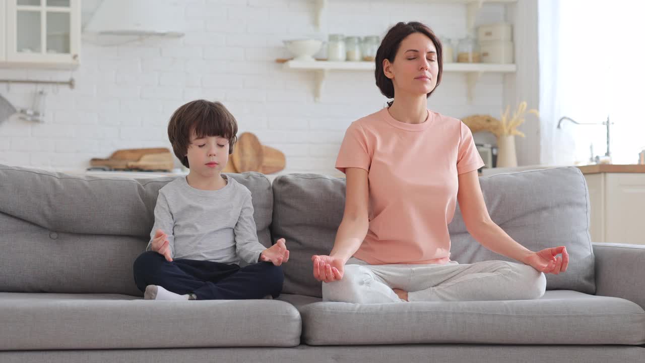 妈妈带着可爱的小儿子一起坐在家里的沙发上练习瑜伽，教孩子冥想视频素材