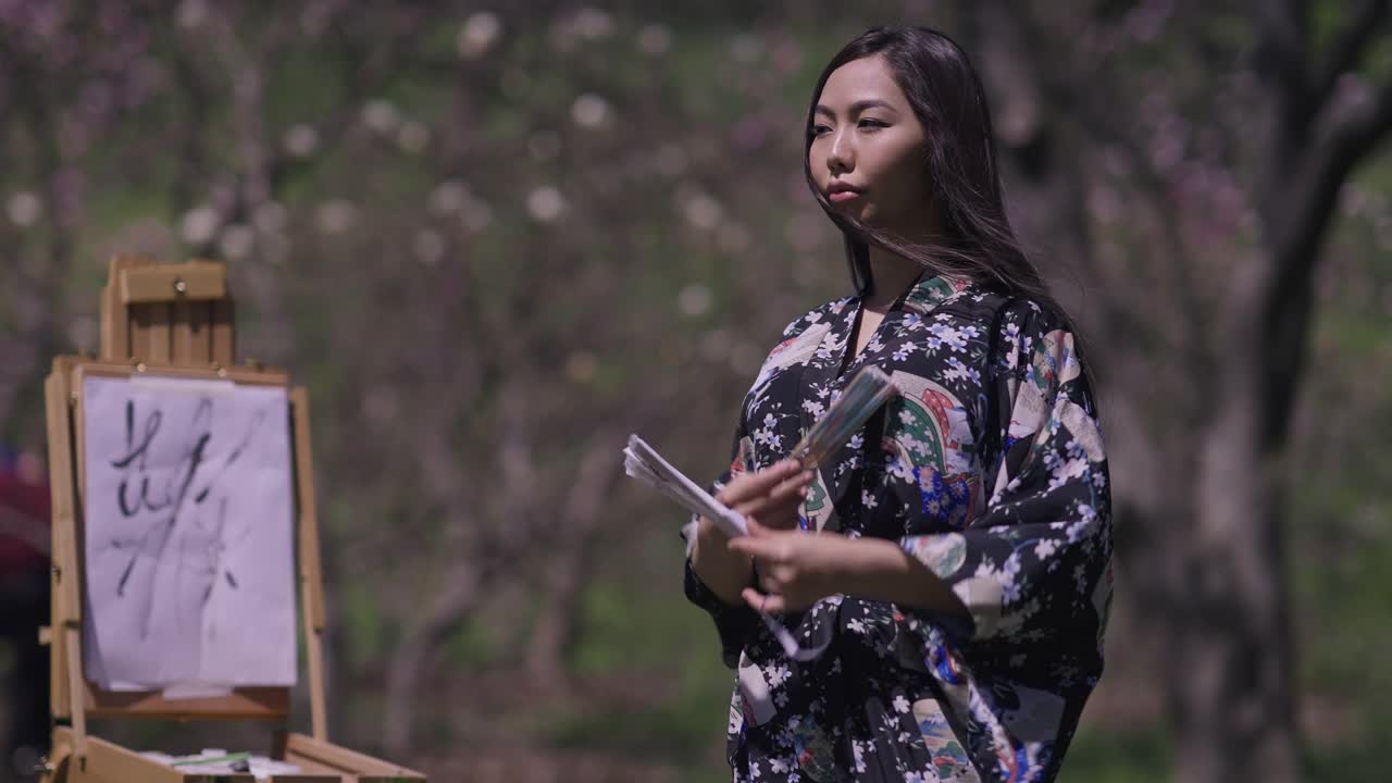 美丽的亚洲年轻女子穿着和服在春天的樱花花园与扇子跳舞微笑与画布画在背景模糊的画架。在阳光明媚的日子里，自信的日本女士的肖像。视频素材