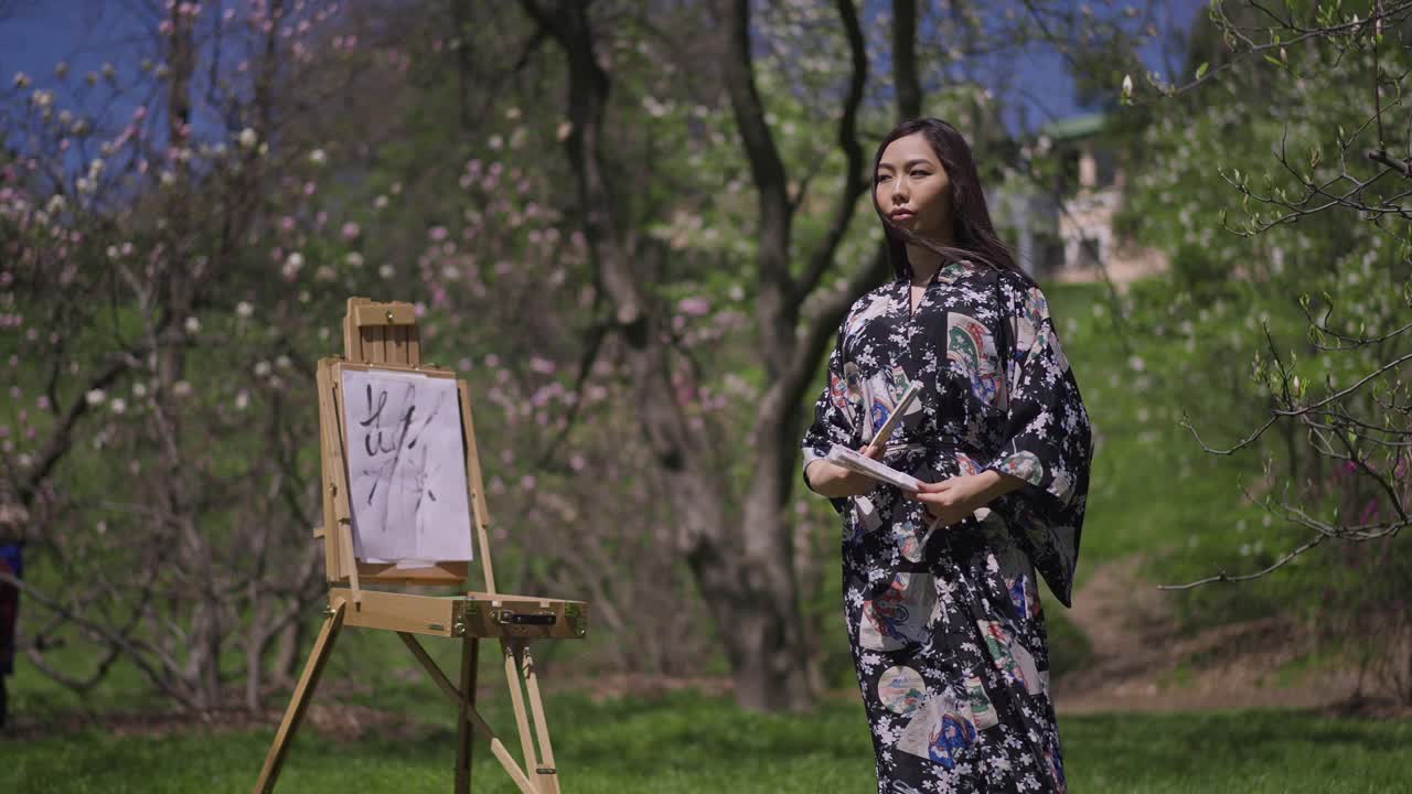 在春暖花开的公园里，一名身穿和服的日本美女在画架前与扇子共舞。艺术的亚洲女士在户外享受阳光的肖像。缓慢的运动。视频素材