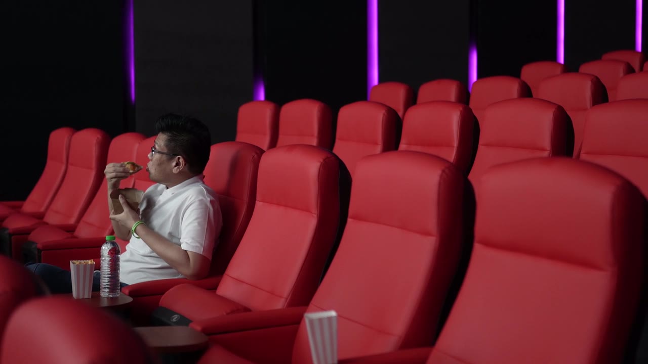 一名亚裔中国男子坐在空荡荡的电影院的红色座位上，一边吃一边看电影。视频素材画面1