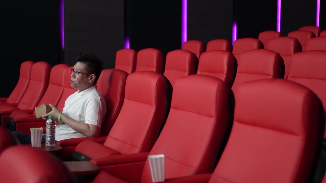 一名亚裔中国男子坐在空荡荡的电影院的红色座位上，一边吃一边看电影。视频素材画面4