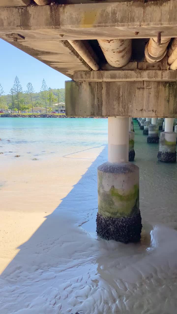 从Tallebudgera Creek桥下走到小溪。下面是牡蛎岩石和沙子。远处的塔勒布杰拉河清澈湛蓝的水。背景是绿树和小山。Tallebudgera Creek澳大利亚昆士兰视频素材