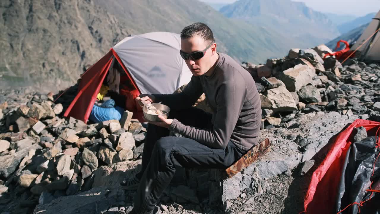 一名男子坐在帐篷旁边的山区营地，吃着铁板粥视频素材