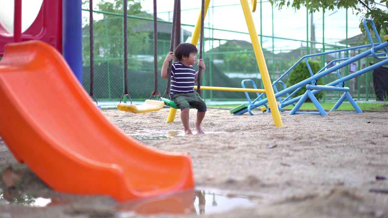 雨后的操场上，一个蹒跚学步的男孩正在玩秋千，他的脚踩在水坑上视频素材