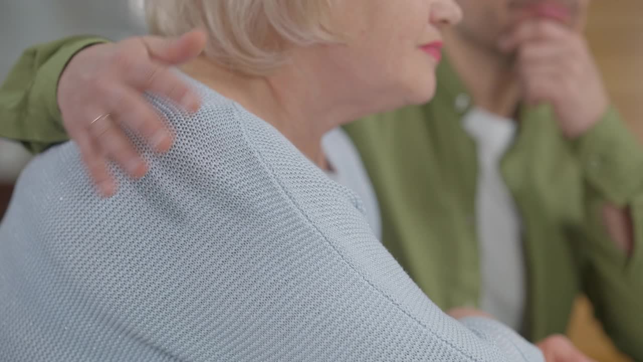 侧视认不出资深女子与年轻男子拥抱抚摩肩。白种人伤心的母亲扶着儿子坐在家里。家庭的悲伤和认可的概念视频素材