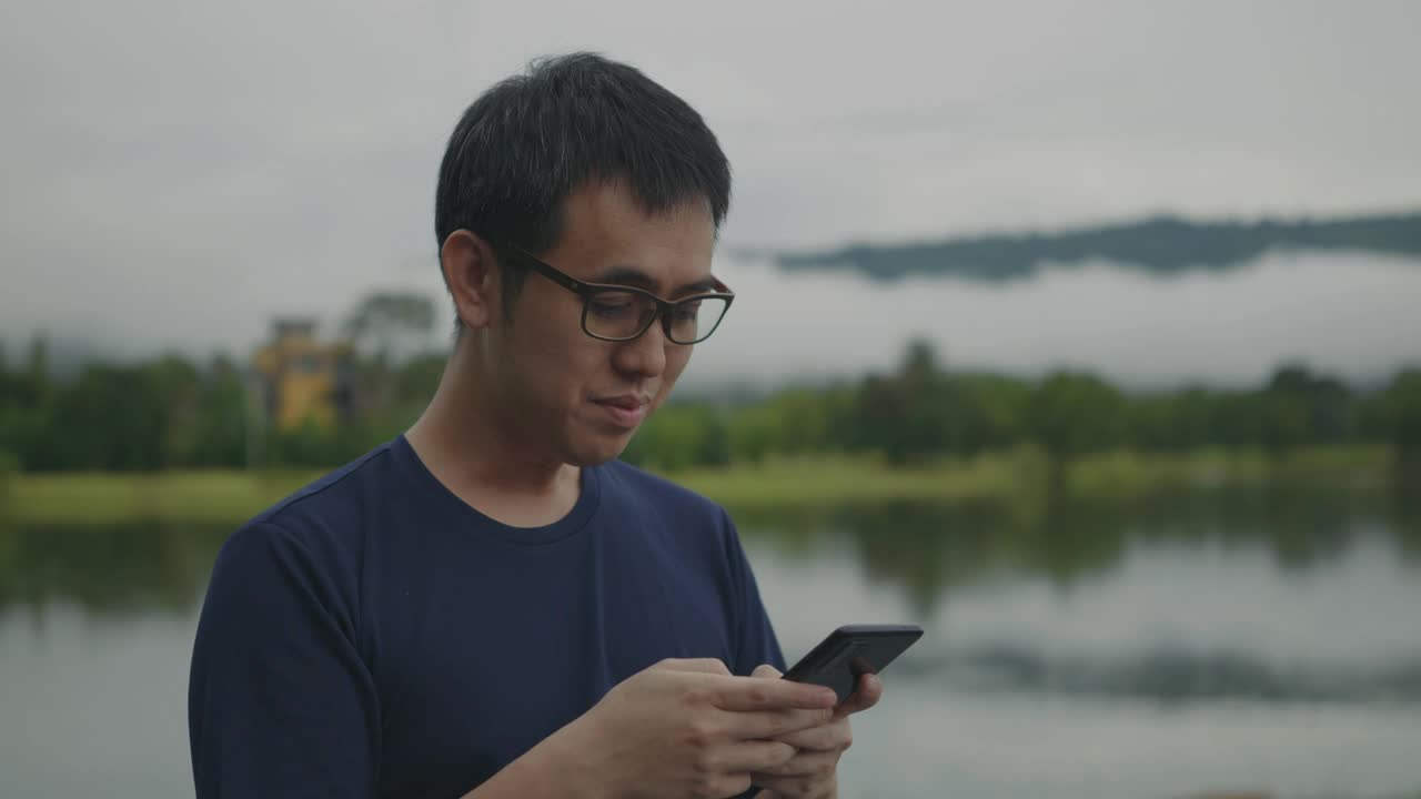 一个亚洲男人在湖边用智能手机看美丽的雨林山景。视频素材