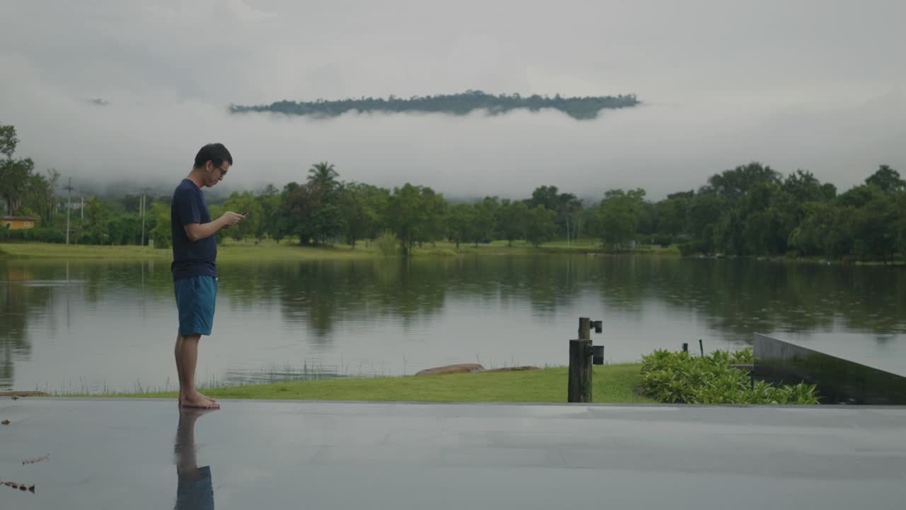 一个亚洲男人在湖边用智能手机看美丽的雨林山景。视频下载