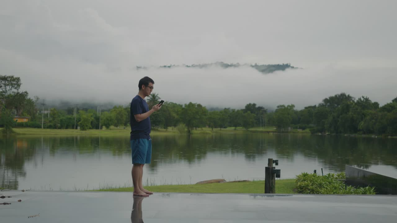 一名亚洲男子正在湖边用智能手机看美丽的雨林山景。视频下载