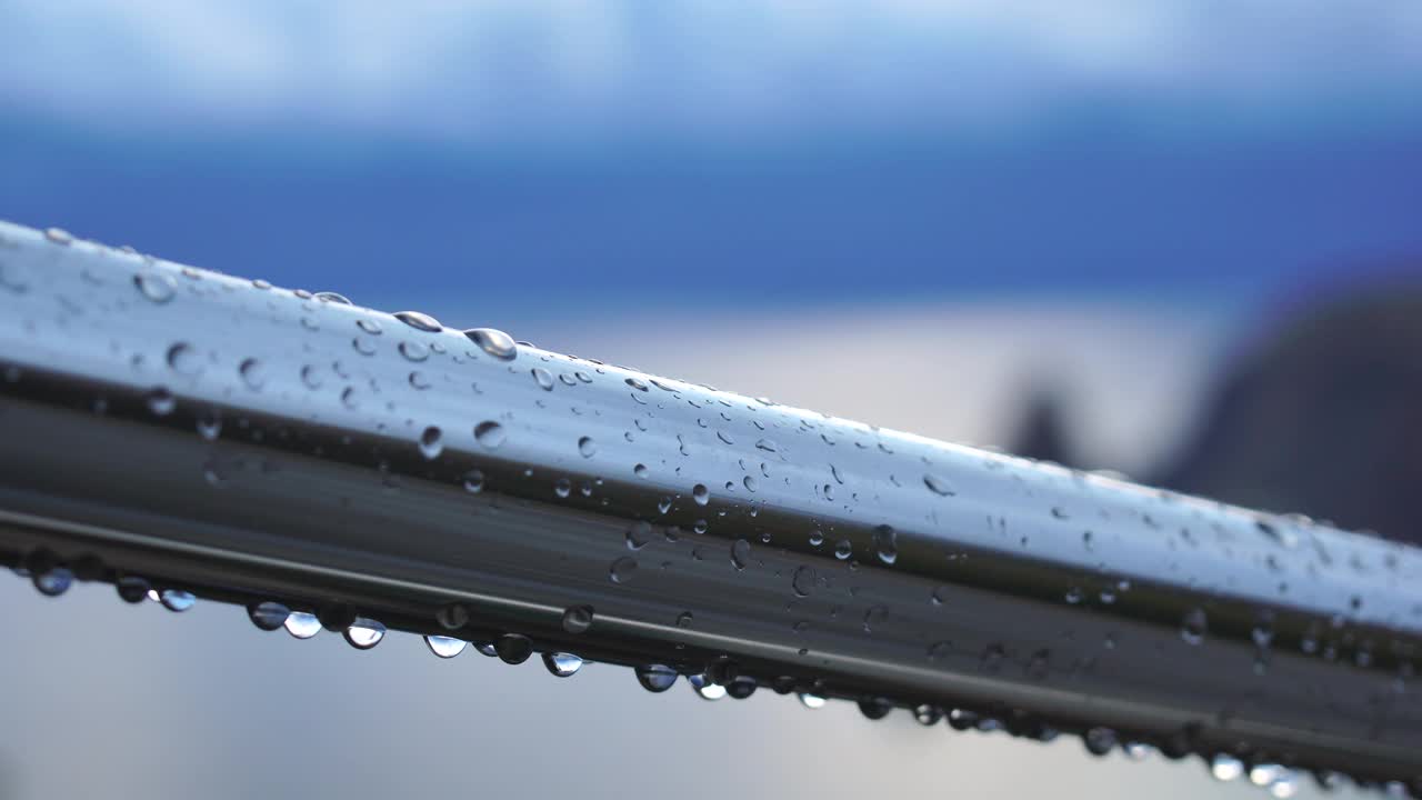 不锈钢扶手上的雨滴映衬着模糊的大海背景。特写镜头。雨露的水滴聚集在闪闪发光的铬钢杆扶手上视频素材