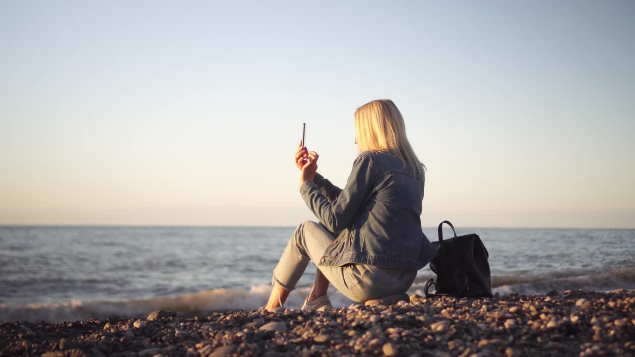 一个女人坐在海边，用手机写着海上的日落。视频素材