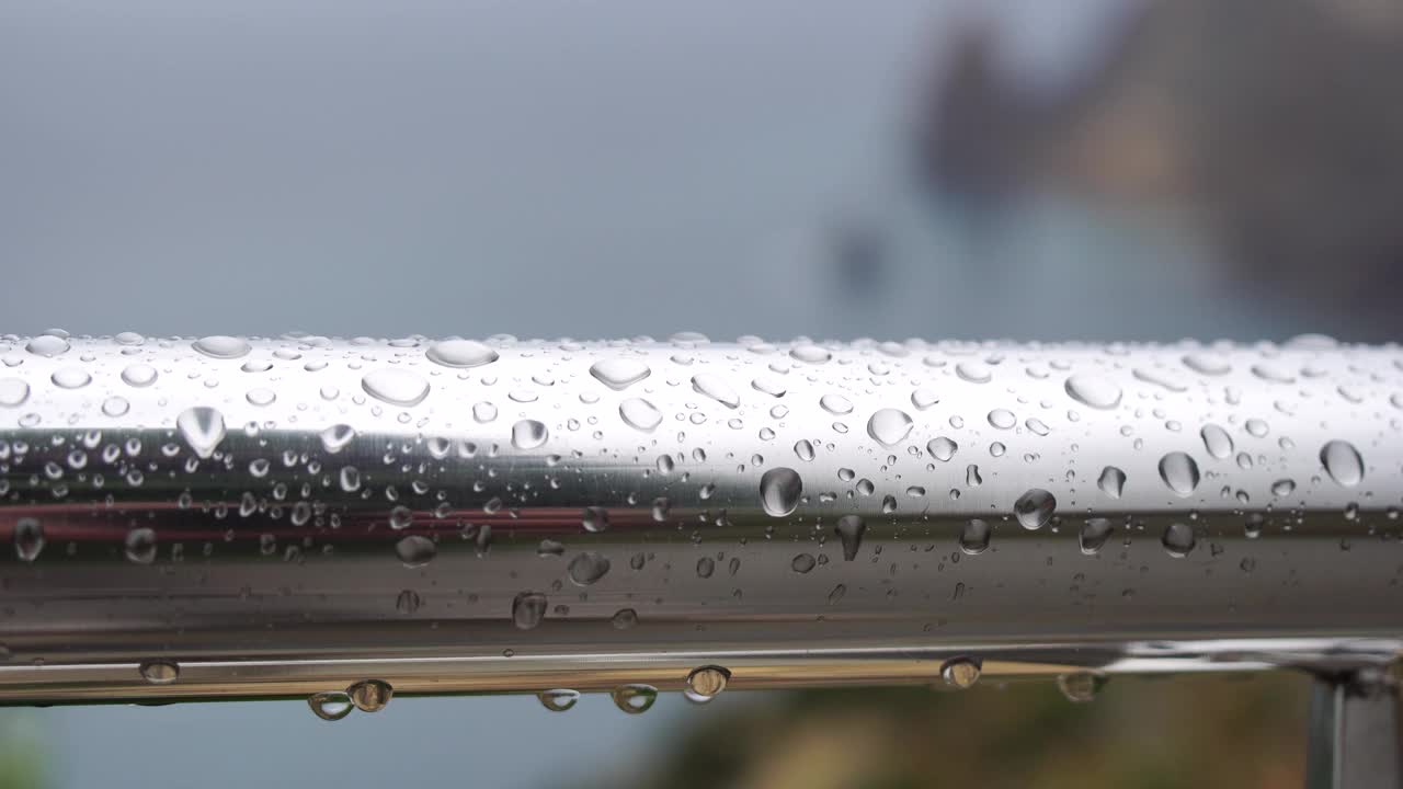 不锈钢扶手上的雨滴映衬着模糊的大海背景。特写镜头。雨露的水滴聚集在闪闪发光的铬钢杆扶手上视频素材