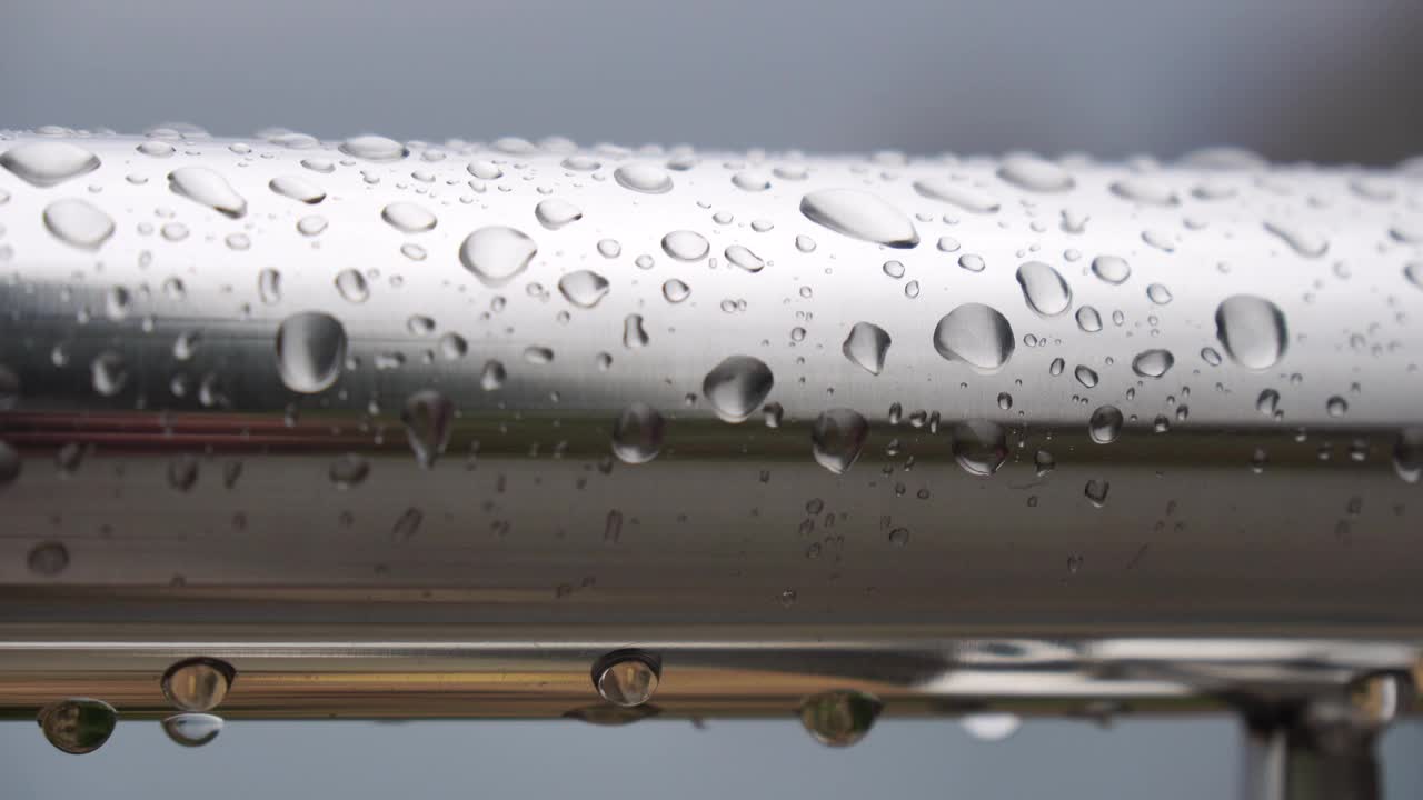 不锈钢扶手上的雨滴映衬着模糊的大海背景。特写镜头。雨露的水滴聚集在闪闪发光的铬钢杆扶手上视频素材