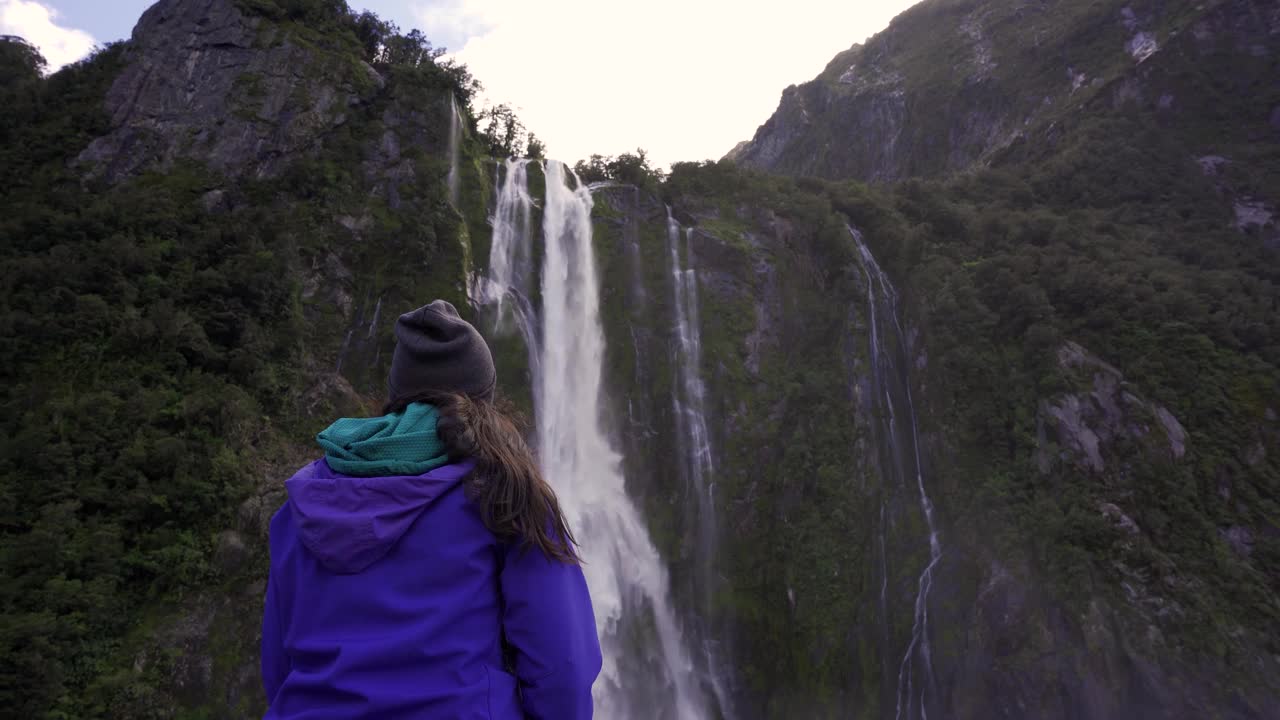 一名年轻女子向后看着米尔福德湾的巨大瀑布。新西兰旅游目的地视频素材