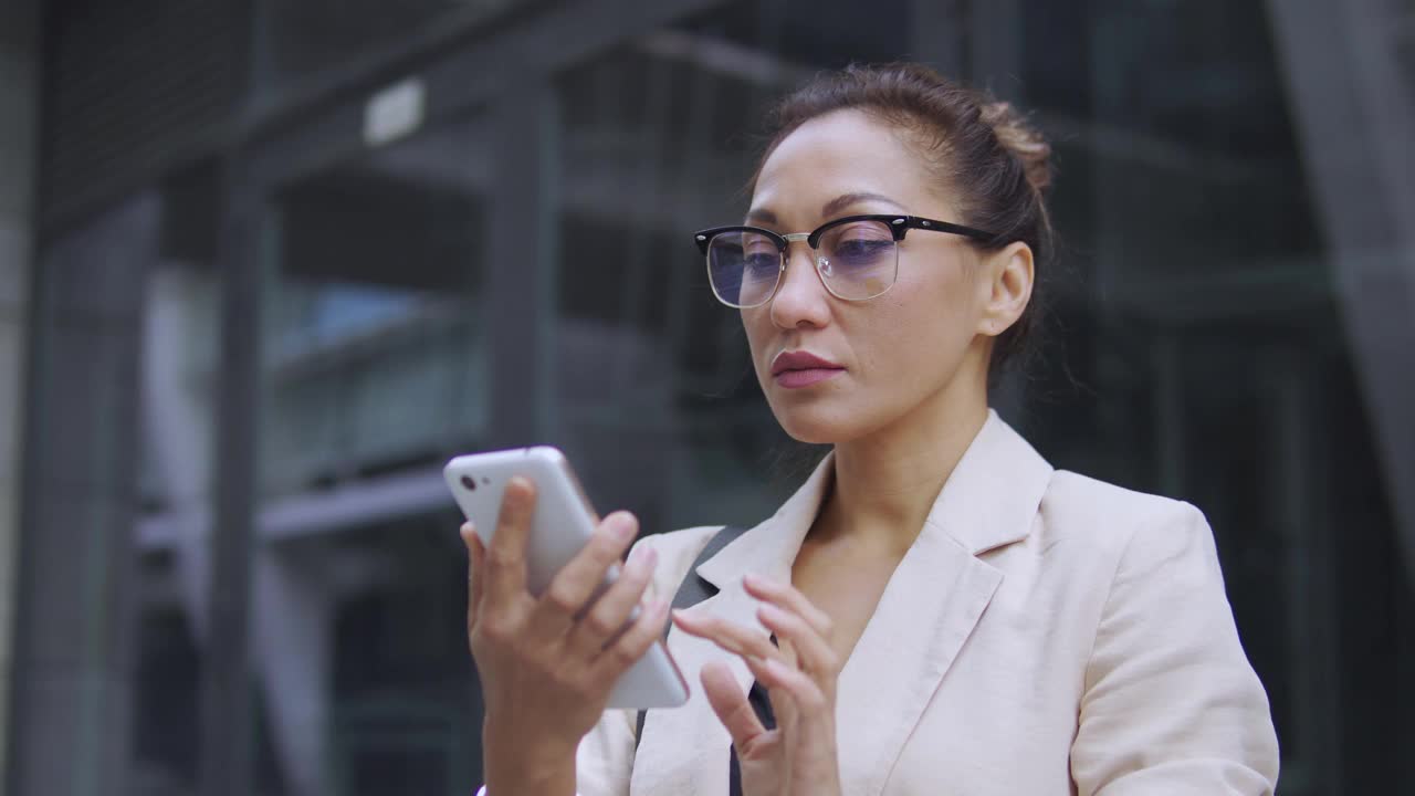 严肃专注的多民族女性戴着眼镜，穿着优雅的西装站在户外用智能手机打字，查收电子邮件回复信件，在群聊中与伙伴聊天，商务生活视频素材