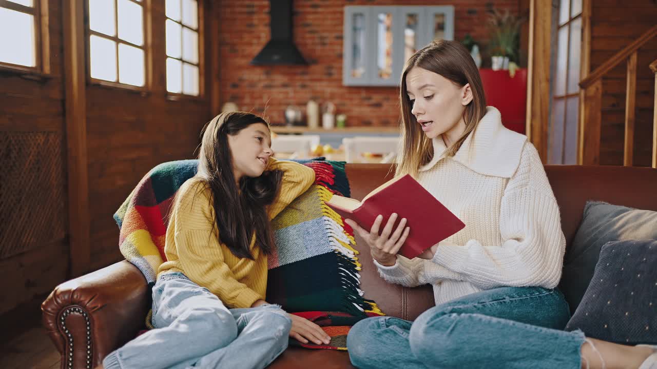 年轻的妈妈抱着书，一边给小女儿读童话，一边坐在木制乡村小屋厨房的沙发上视频素材