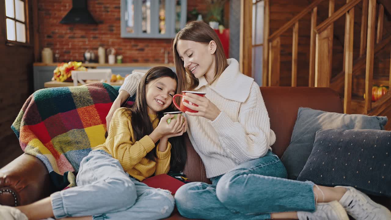 年轻的母亲和小女儿端着热饮，微笑着拥抱着。坐在家里厨房的沙发上。特写镜头，慢镜头视频素材