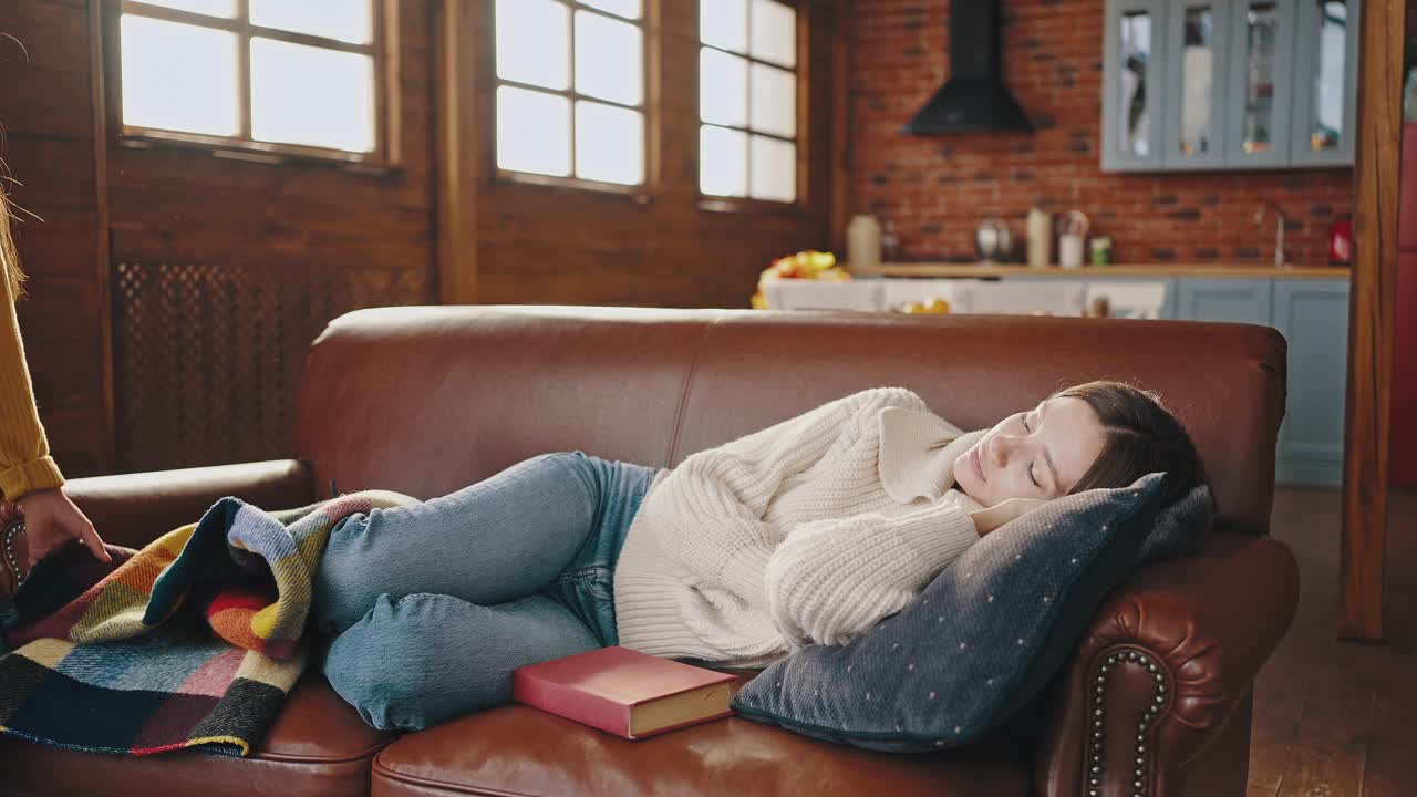 年轻的母亲在乡村别墅厨房的沙发上拿着书睡着了。小女儿向她走来，用格子呢盖住妈妈，拥抱着她视频素材