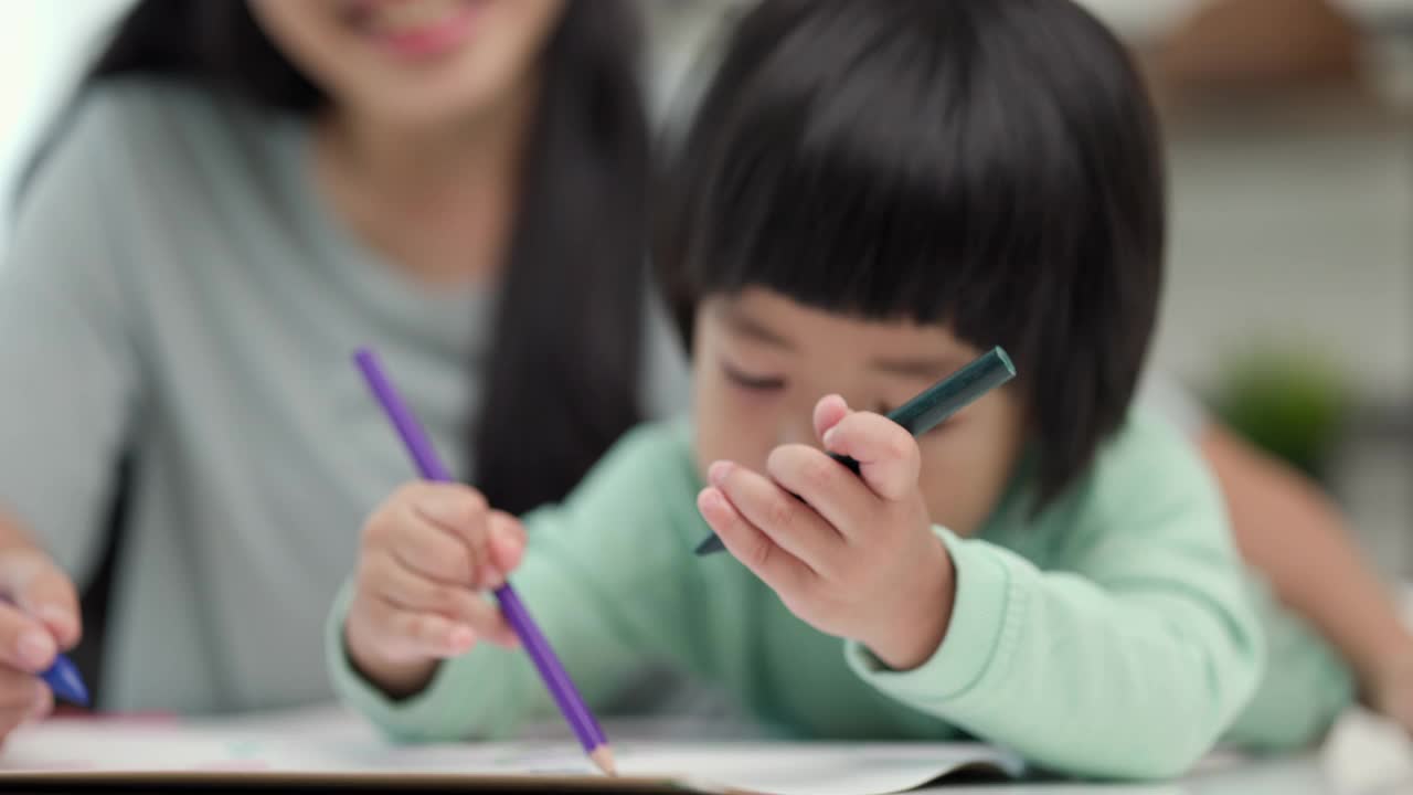 快乐的亚洲男孩与他的母亲在客厅里用蜡笔和彩色铅笔画画。妈妈教儿子如何用蜡笔在书上画画或做作业。家庭的概念。视频素材