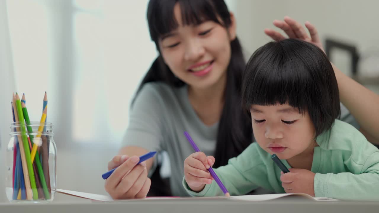 快乐的亚洲男孩与他的母亲在客厅里用蜡笔和彩色铅笔画画。妈妈教儿子如何用蜡笔在书上画画或做作业。家庭的概念。视频素材