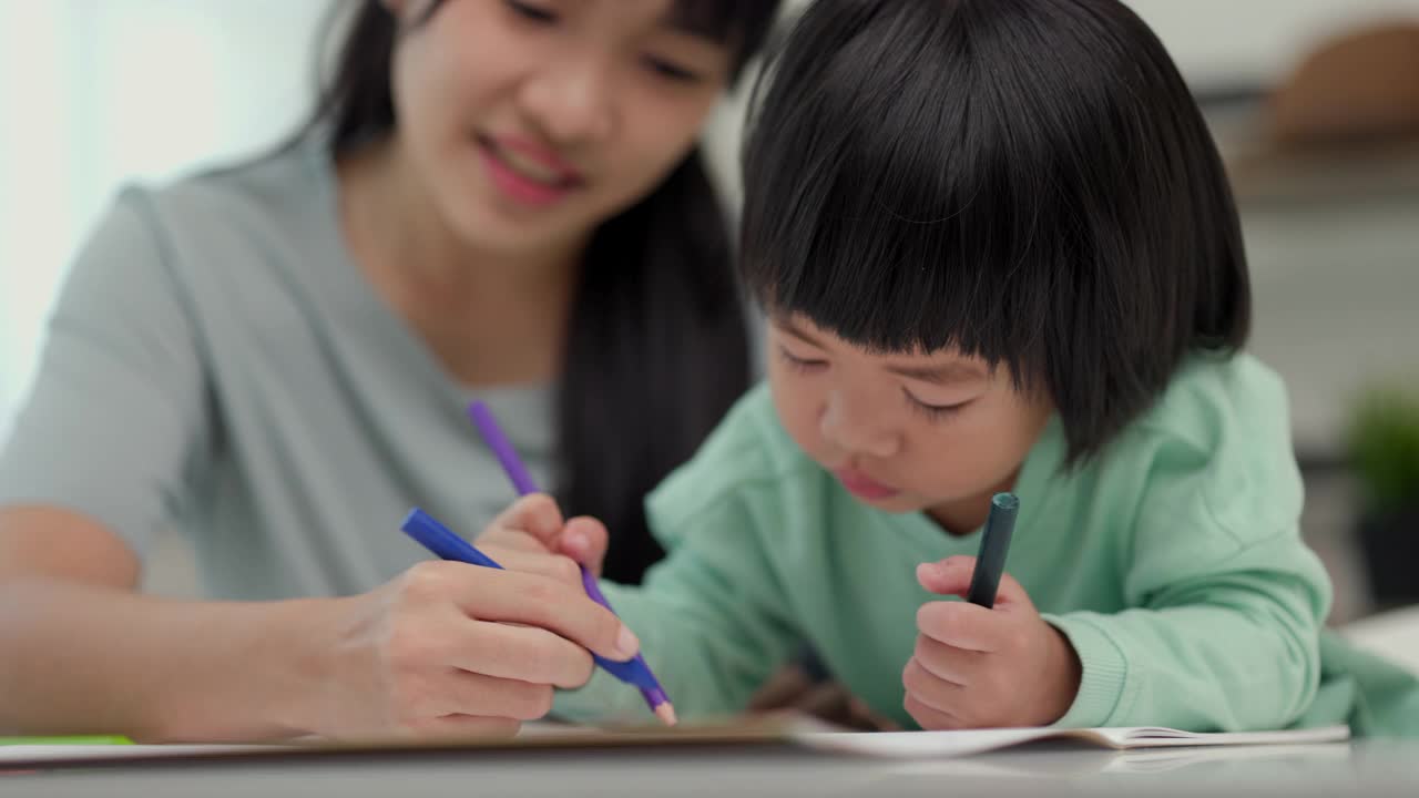 快乐的亚洲男孩与他的母亲在客厅里用蜡笔和彩色铅笔画画。妈妈教儿子如何用蜡笔在书上画画或做作业。家庭的概念。视频素材