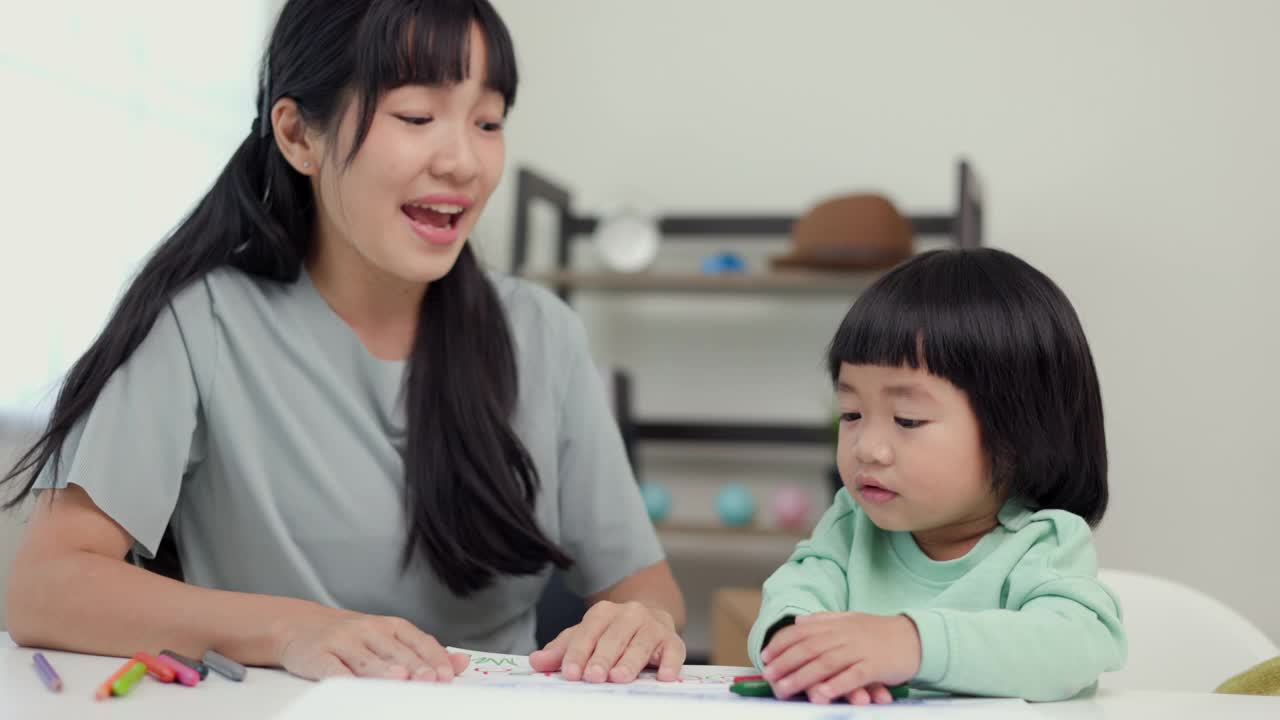 快乐的亚洲男孩与他的母亲在客厅里用蜡笔和彩色铅笔画画。妈妈教儿子如何用蜡笔在书上画画或做作业。家庭的概念。视频素材