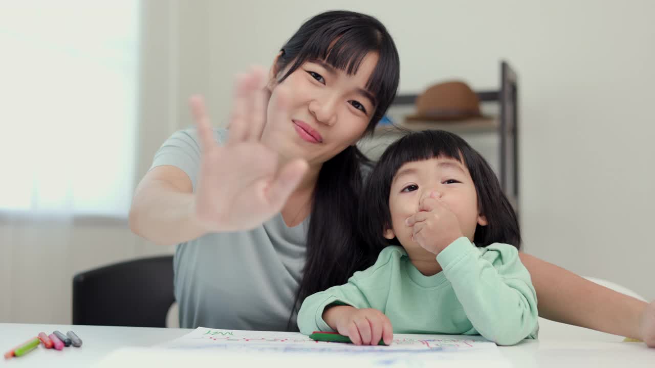 快乐时刻亚洲妈妈和小男孩在家里用蜡笔画画的时候对着镜头说你好或再见。家人在房子里玩得开心。妈妈教儿子做作业。视频素材