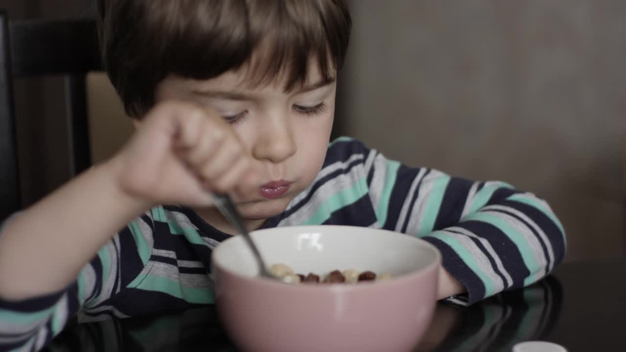 孩子吃牛奶玉米片。小饥饿的男孩坐在厨房的桌子上吃着早餐的玉米麦片。健康的营养片。孩子的肖像吃早餐吃什锦麦片视频素材