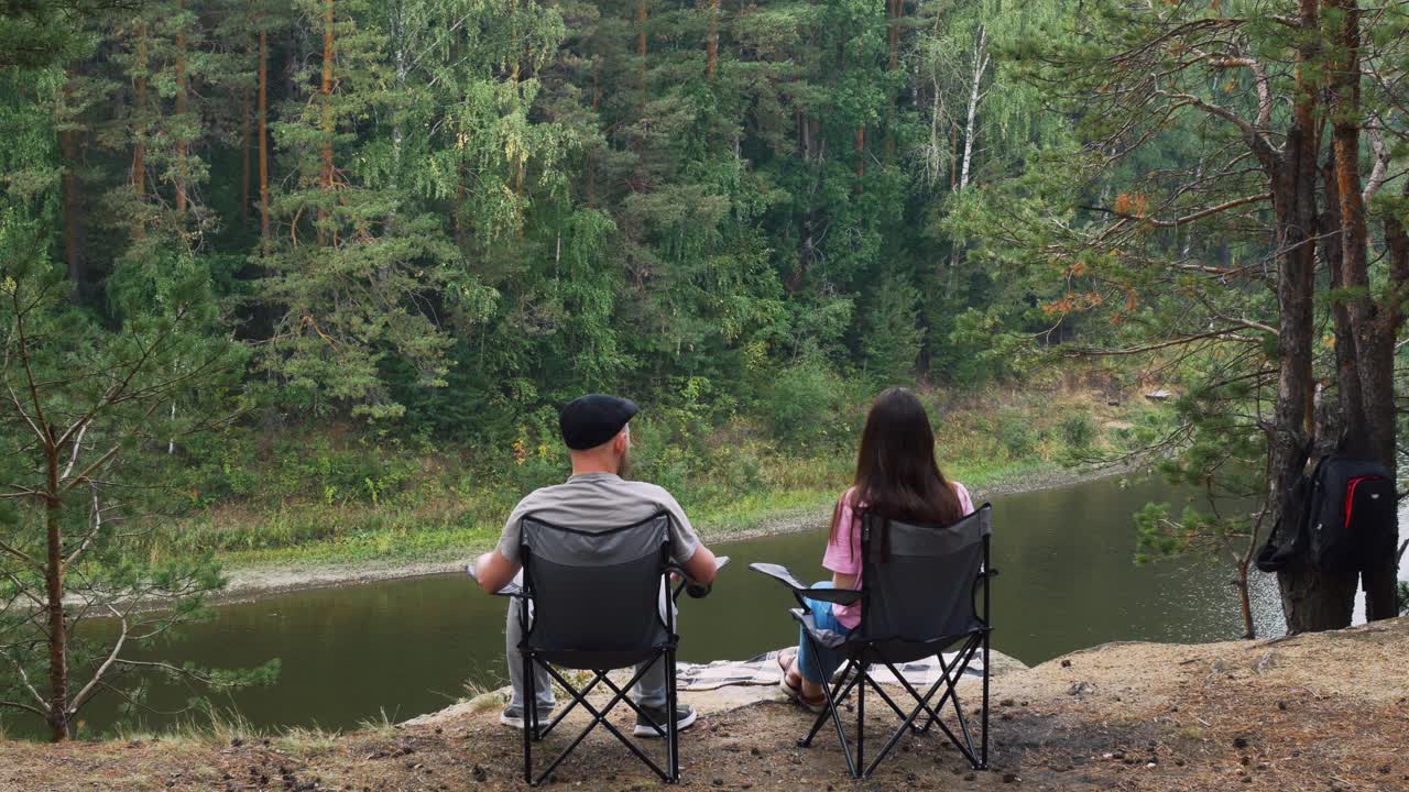 野营时，一男一女手牵着手坐在户外的扶手椅上，欣赏着森林和山河的美丽景色。从工作周中休息在自然中。从后面看视频素材