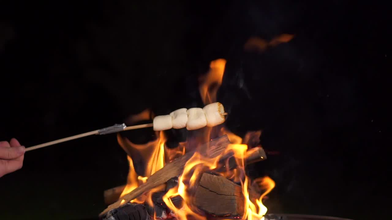 棉花糖棒烤篝火在黑色背景。人们在壁炉旁烤棉花糖，享受他们的假期。朋友们在烤架上烤棉花糖的特写。视频素材