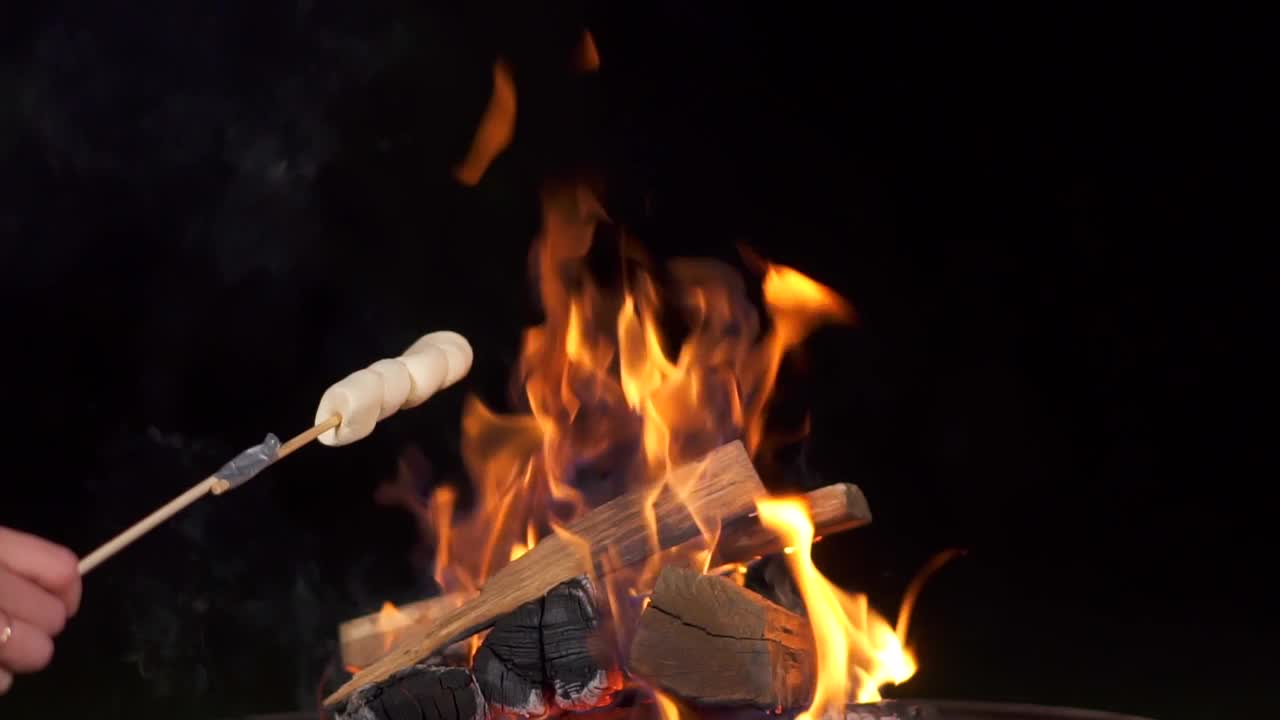 棉花糖棒烤篝火在黑色背景。人们在壁炉旁烤棉花糖，享受他们的假期。朋友们在烤架上烤棉花糖的特写。视频素材