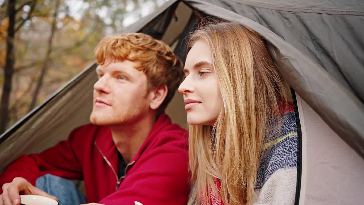 年轻女子把头靠在坐在帐篷里端着杯茶微笑的男友肩上。森林里的秋日。近距离视频素材