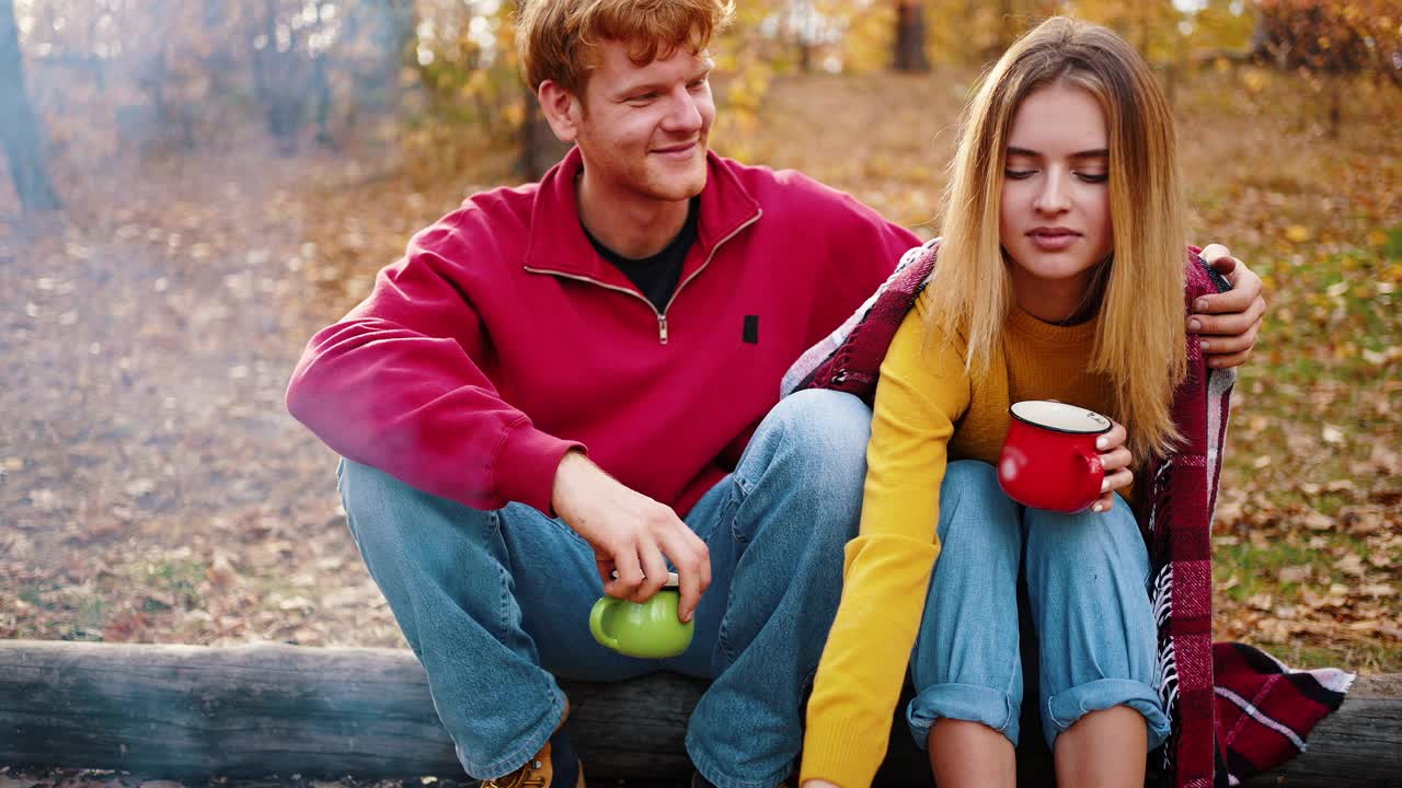 年轻男子拥抱女友谁拿着杯茶而坐在原木在秋天的木材。野餐,篝火的烟雾。特写，慢动作视频素材