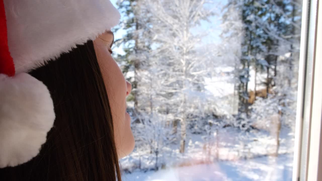 戴着圣诞老人红帽的无忧无虑开朗的千禧妇女坐在窗台上看着白雪美丽平静的森林，在家享受庆祝新年、圣诞、寒假的喜庆心情视频素材