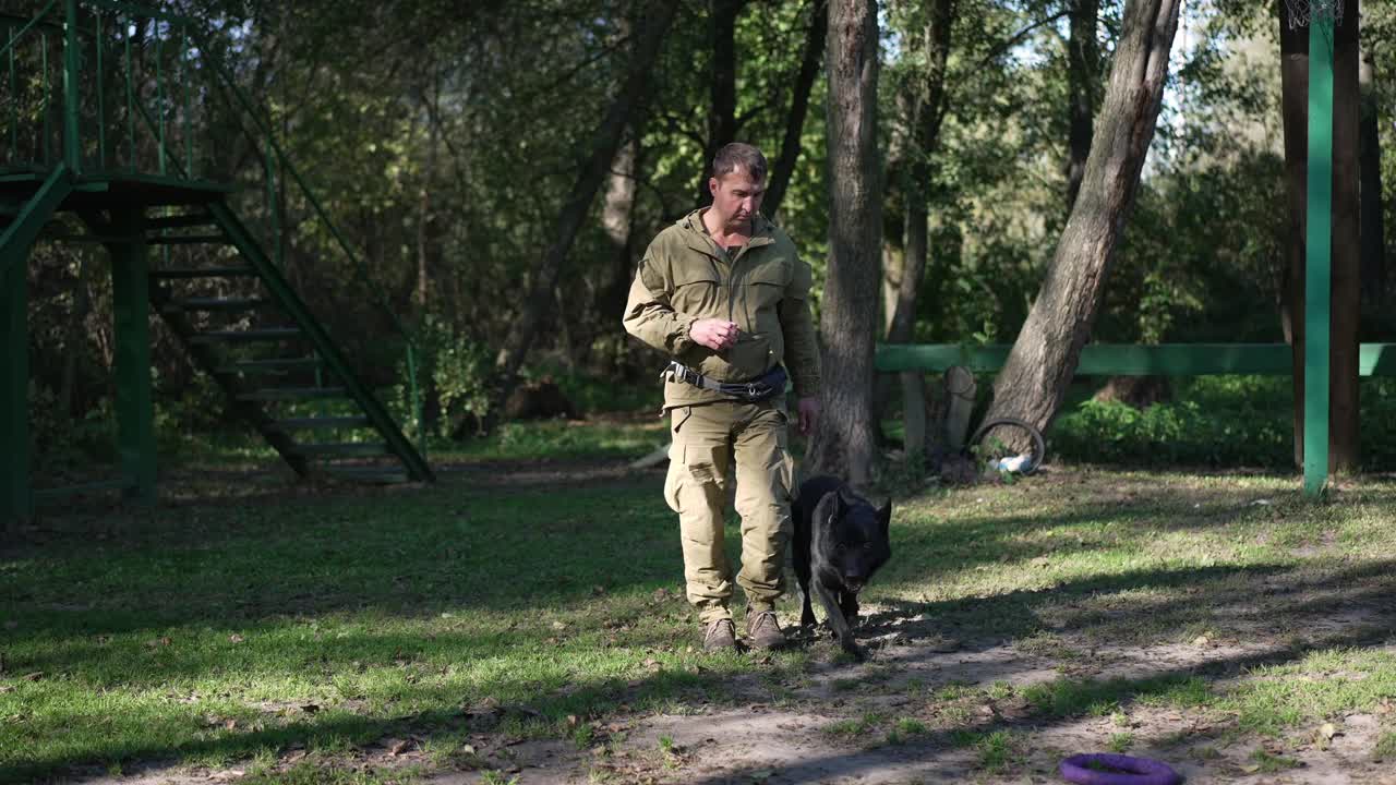 严肃的专业犬类专家慢动作地走着，华丽的黑色德国牧羊犬跟着驯兽师踱步。阳光明媚的户外，温顺的狗和自信的人。视频素材