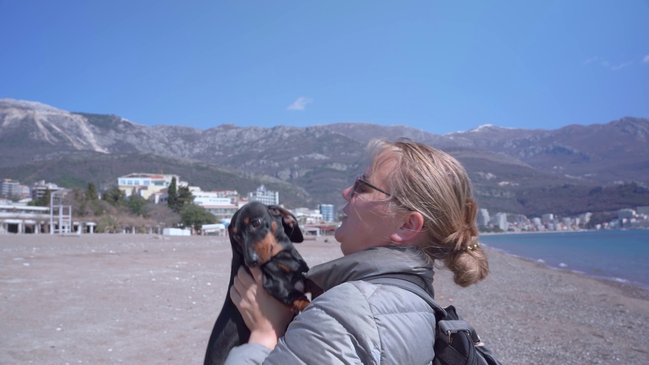 年轻的金发女游客带着她的腊肠犬在海边散步，把小狗扔起来，亲吻它。和宠物一起在海边度假视频素材