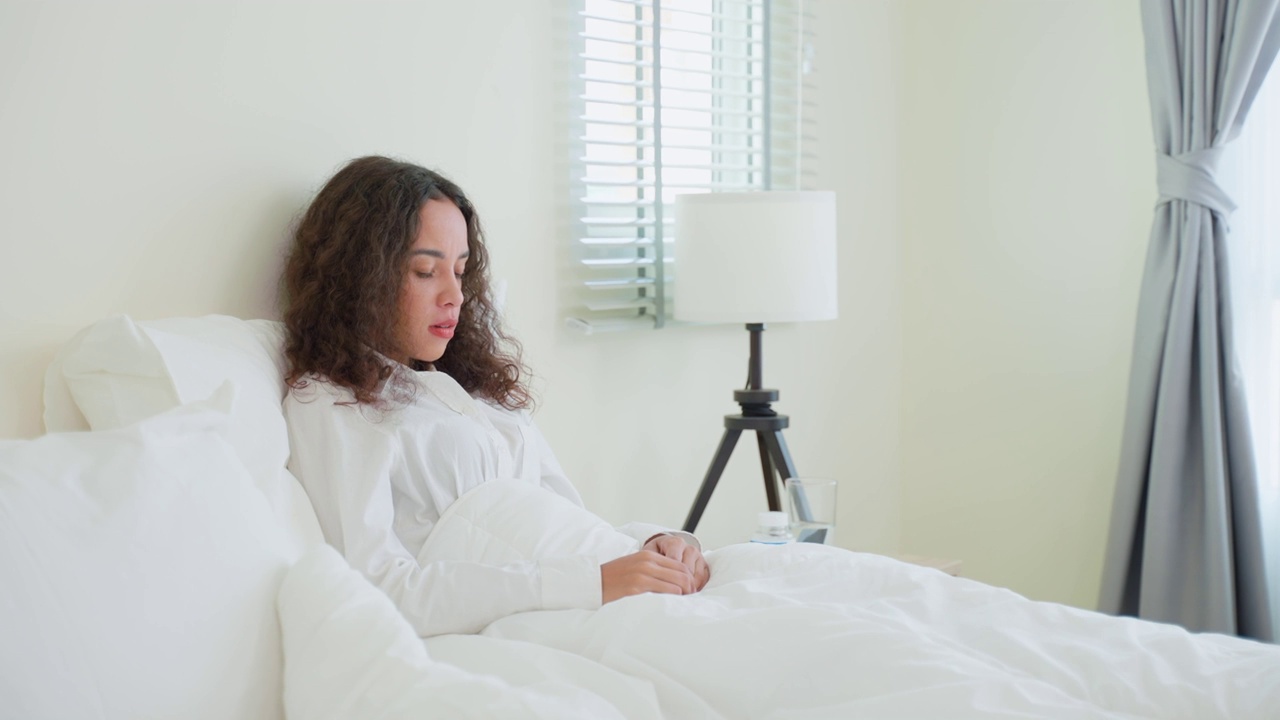 美丽的拉丁病号女孩穿着睡衣从卧室里醒来。漂亮的年轻女子生病感觉不好，醒来后在床上咳嗽，发烧或感冒病毒在早上在家。视频素材