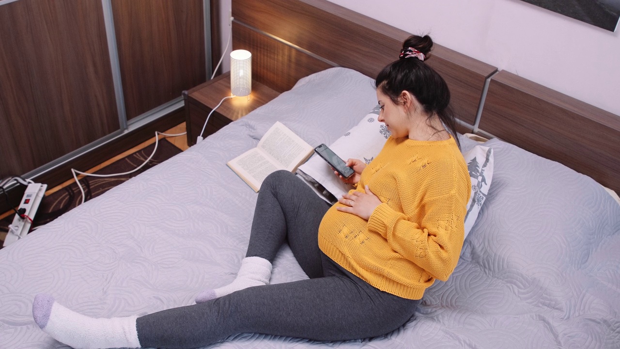 孕妇一边在卧室看书一边上网。视频素材画面1