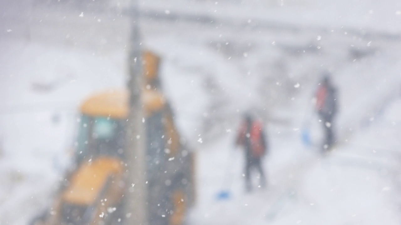 在冬天的暴风雪中清扫街道。工人们拿着铁锹和带着铲斗的拖拉机在离焦的道路上清理积雪。选择性聚焦，浅景深。大雪视频素材