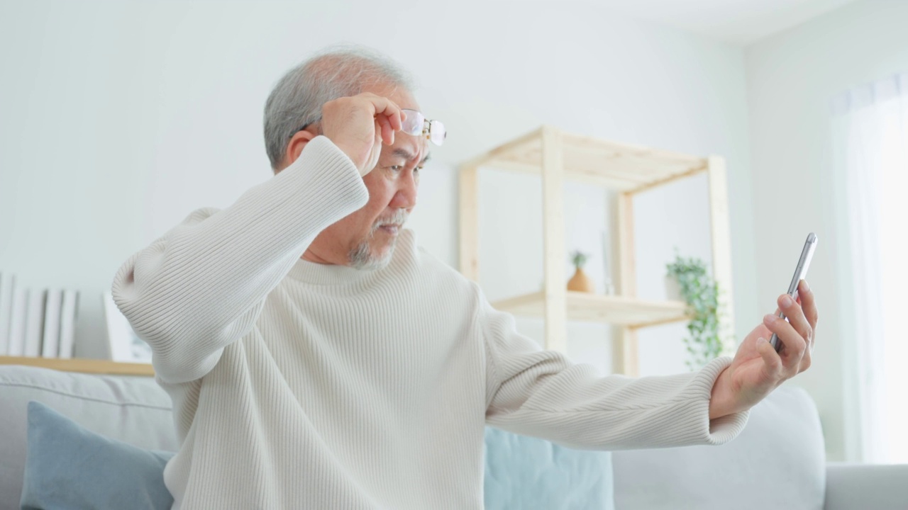 在家里客厅使用手机的亚洲老年男性。强壮年长成熟的爷爷戴着眼镜，坐在沙发上用智能手机聊天，与家人交流，享受退休生活。视频素材