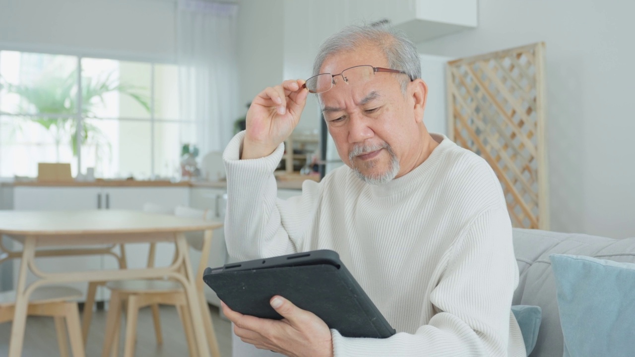 亚洲魅力老人在家客厅使用平板电脑。年长成熟的爷爷戴着眼镜坐在沙发上，用智能手机聊天，与家人交流，享受退休生活。视频素材