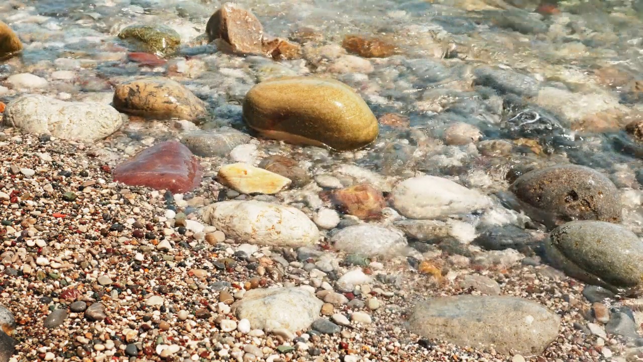 阳光明媚的日子里，布满卵石的海滩上的水边。沙滩上五颜六色的鹅卵石，清澈透明的水波。岩石和石头的蓝绿色的大海。石海滩。视频素材