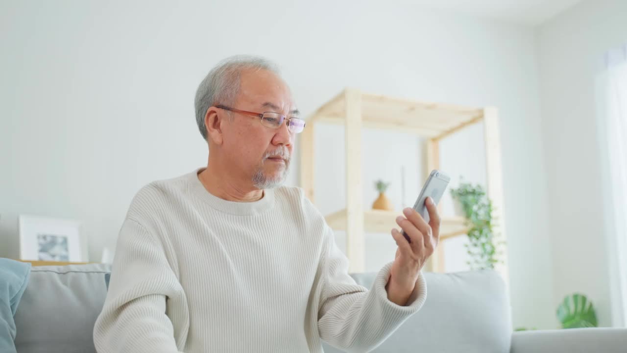 在家里客厅使用手机的亚洲老年男性。强壮年长成熟的爷爷戴着眼镜，坐在沙发上用智能手机聊天，与家人交流，享受退休生活。视频素材