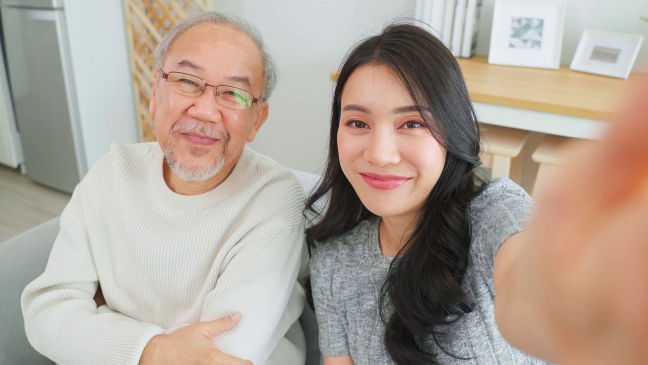 亚洲可爱的家庭，女儿用电话与资深父亲视频通话。年轻漂亮的女孩和上了年纪的老男人坐在沙发上享受网上交流，在家里的客厅里一起聊天视频素材