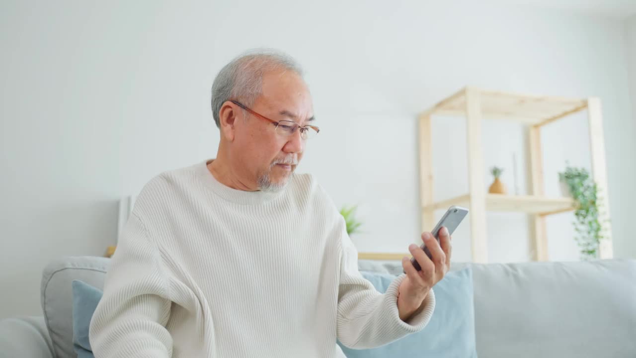 在家里客厅使用手机的亚洲老年男性。强壮年长成熟的爷爷戴着眼镜，坐在沙发上用智能手机聊天，与家人交流，享受退休生活。视频素材