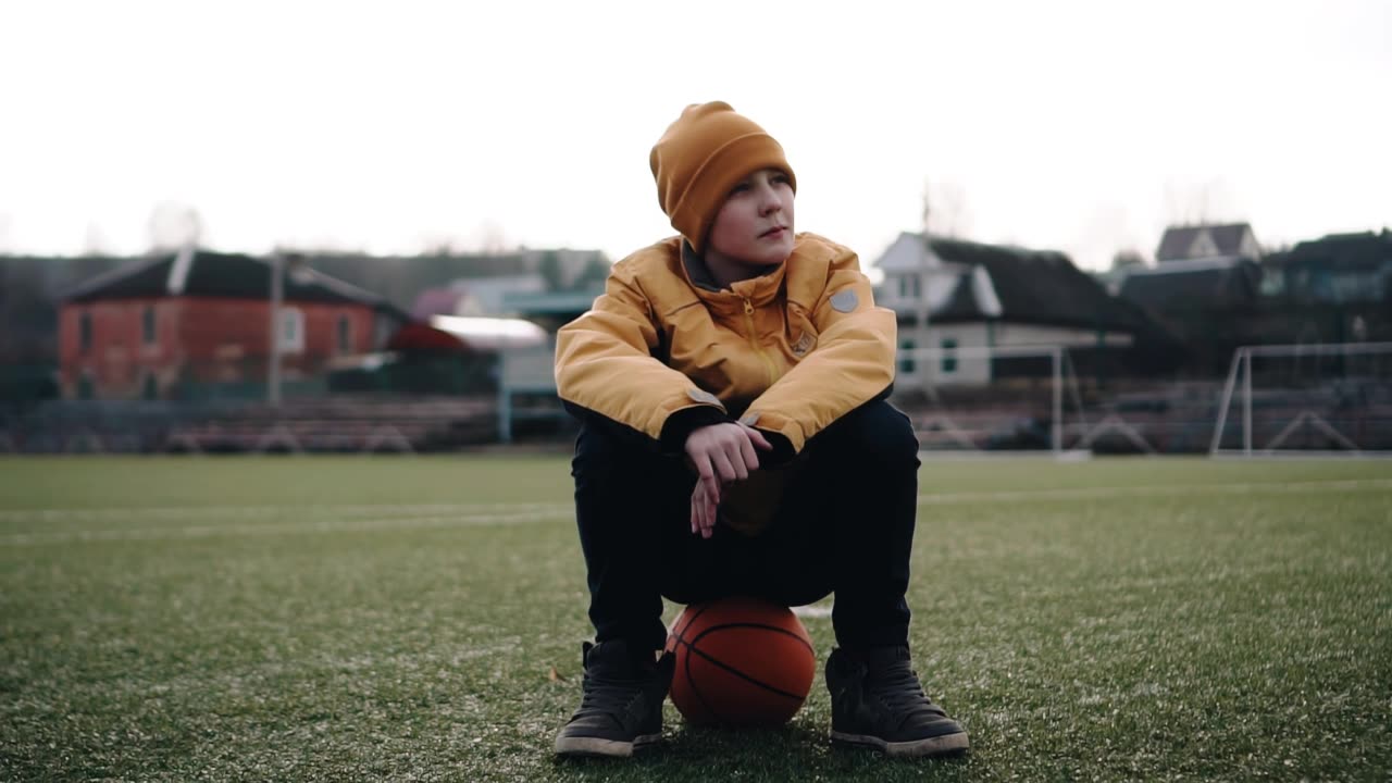 一个男孩坐在体育场草地上的球上。他望向远方视频素材画面1