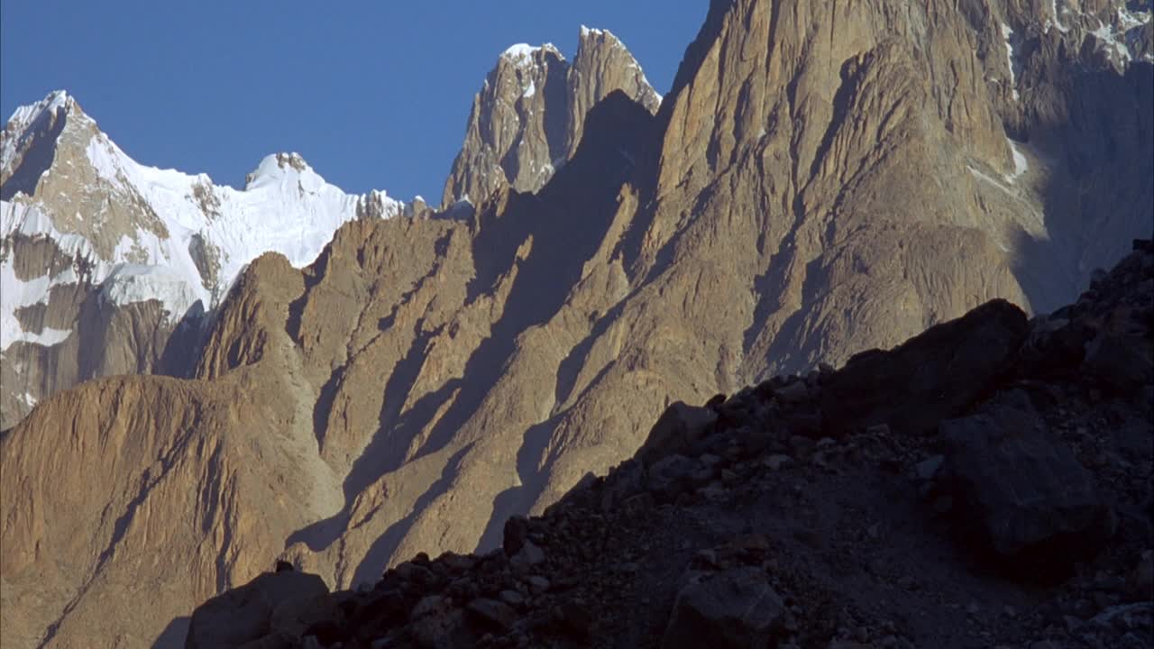 三个登山者爬上陡峭的岩石斜坡的跟踪镜头。登山者在山的阴影里。在背景中看到金字塔形状的山峰。山峰被雪覆盖着。视频素材